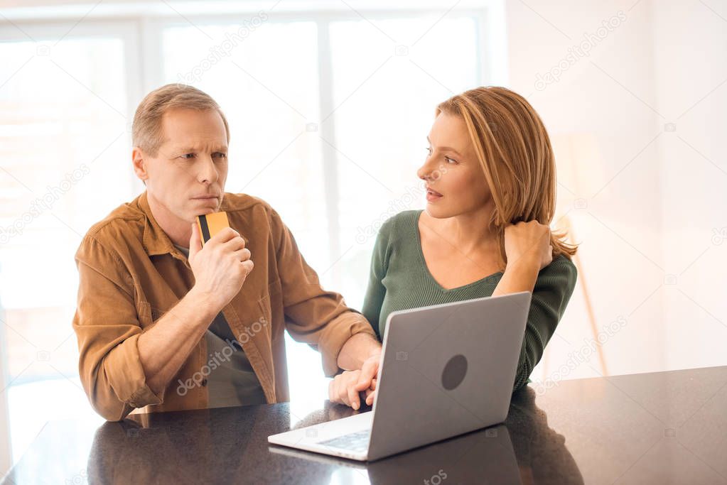 thoughtful man holding credit card while using laptop together with wife