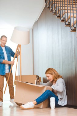 smiling man holding floor lamp while woman sitting in floor and unpacking carton box clipart