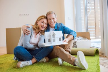 happy couple hugging and demonstrating house model while sitting on floor on green carpet clipart