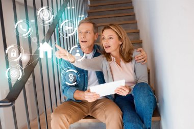 wife pointing hand while sitting with husband on stairs and using digital tablet, smart home concept clipart