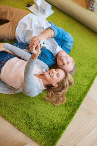 selective focus of woman holding keys with house model trinket while lying on green carpet with smiling husband