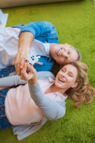 selective focus of happy couple holding keys with house model trinket while lying on green carpet 