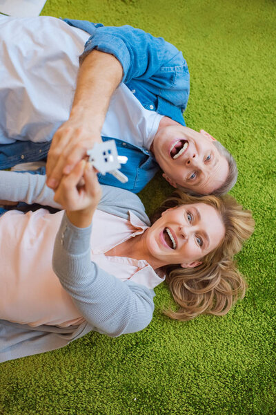 selective focus of excited couple holding keys with house model trinket while lying on green carpet 