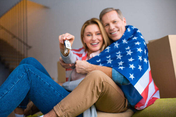selective focus of woman holding keys with house model trinket while wrapping in usa national flag with husband