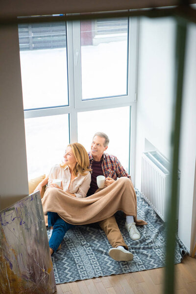 happy couple sitting on floor by large window and holding coffee cups
