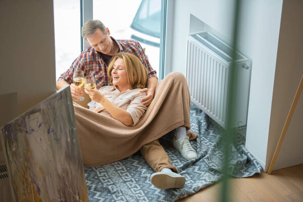 happy couple clinking glasses while resting on floor by large window at new home