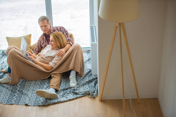 happy couple using laptop while resting on floor by large window at new home
