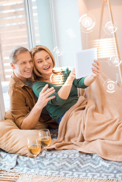 smiling couple using digital tablet while sitting under fleece blanket on floor, smart home concept
