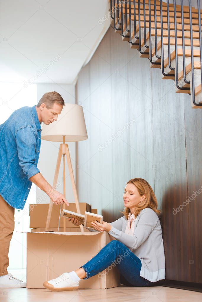 woman sitting on floor and unpacking cardboard box and man pointing with finger