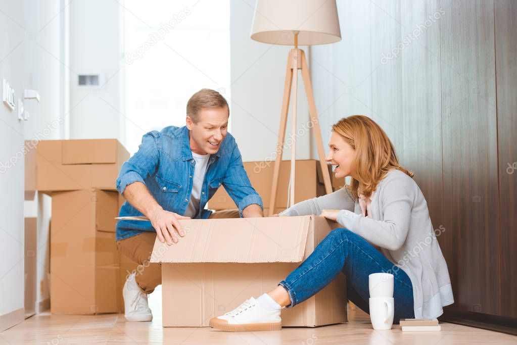 cheerful couple sitting on floor and unpacking cardboard box