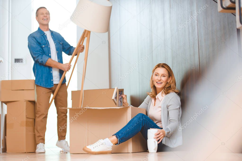 selective focus of woman sitting on floor and unpacking cardboard box while husband holding floor lamp