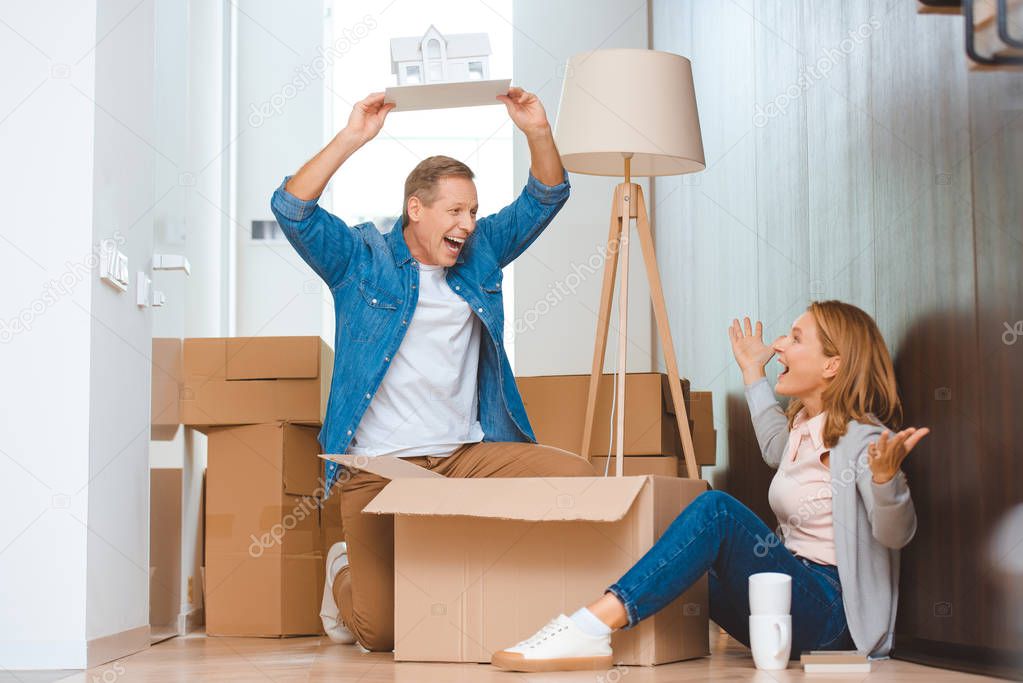 excited man holding house model over head while happy sitting on floor near cardboard box