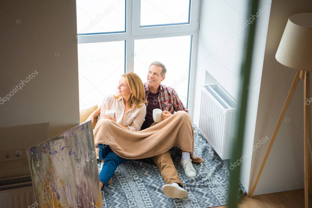 smiling couple with coffee cups sitting on floor by large window at new home