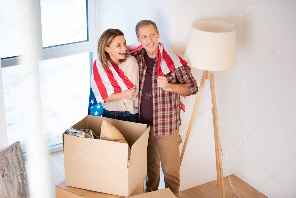 selective focus of happy couple wrapping in usa national flag while standing near cardboard boxes at new home