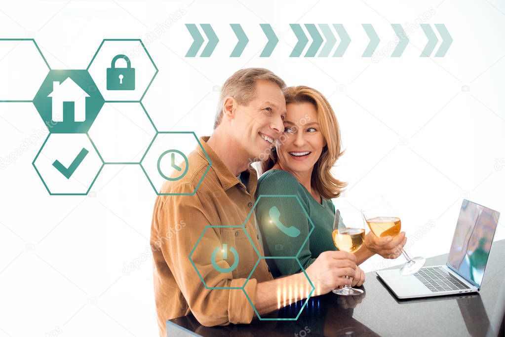 happy couple holding glasses of white wine while standing by table with laptop, smart home concept