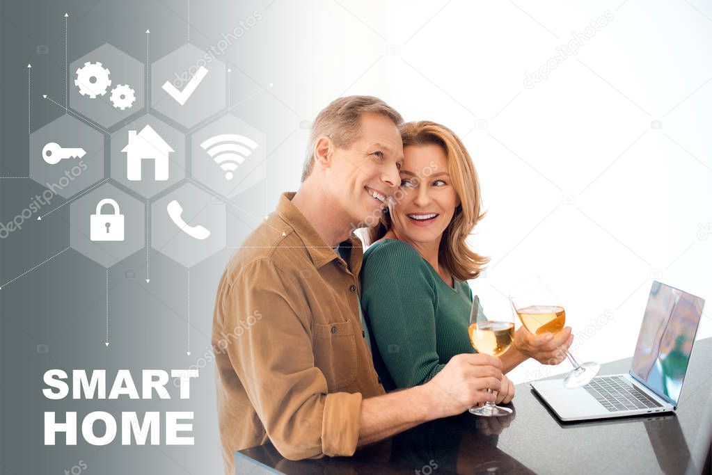 happy couple holding glasses of white wine while standing by table with laptop, smart home concept