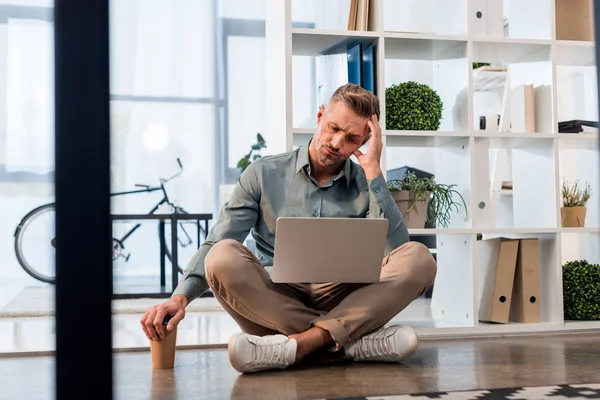 handsome businessman sitting with crossed legs on floor and looking at laptop
