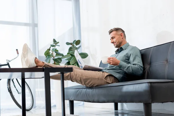 Alegre Hombre Negocios Sentado Sofá Sosteniendo Taza Mientras Lee Periódico — Foto de Stock