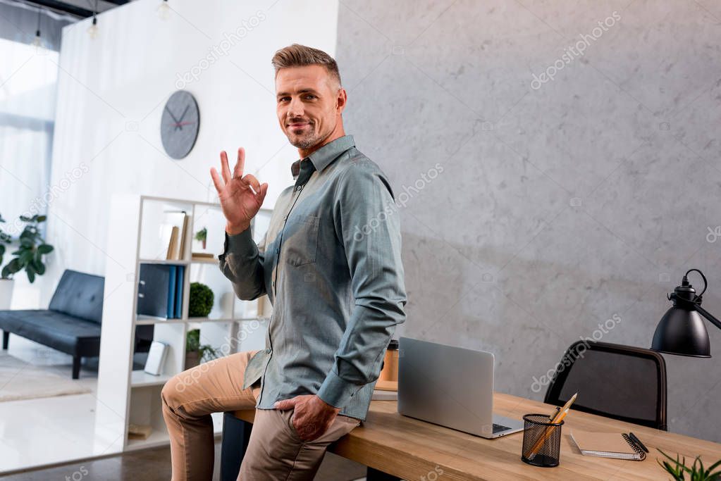 happy businessman showing ok sign while sitting with hand in pocket in office 