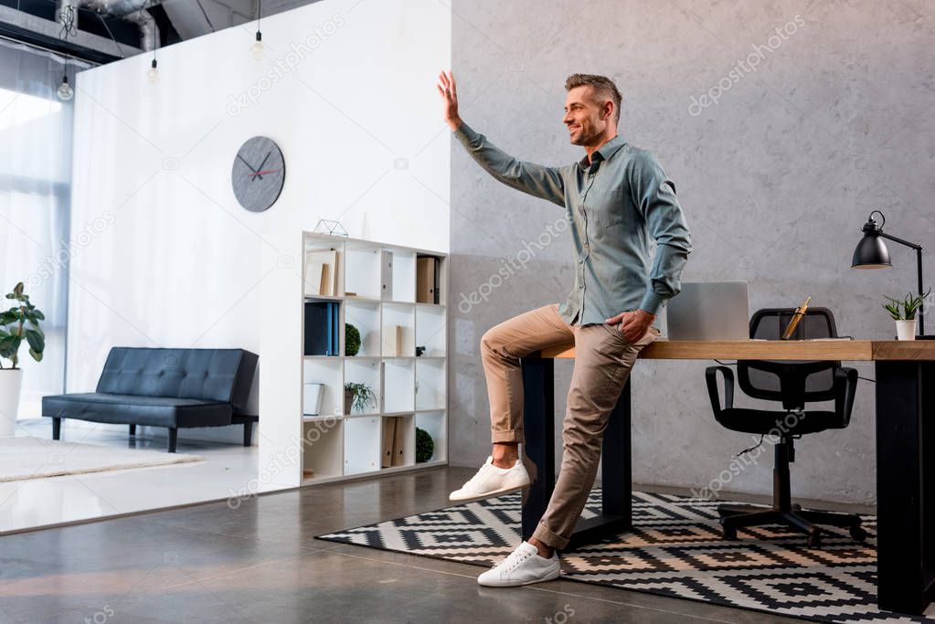 cheerful businessman waving hand and standing with hand in pocket in modern office