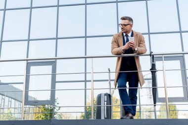 bavul ve şemsiye yakın dururken smartphone kullanarak bej bir palto neşeli işadamı 