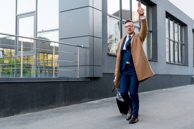 bej bir palto bavul ve şemsiye ile sokakta çalışırken barış işareti gösterilen işadamı