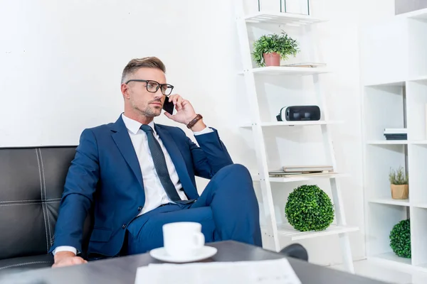 在智能手机上交谈和坐在咖啡桌附近的沙发上的英俊商人的选择性焦点 — 图库照片