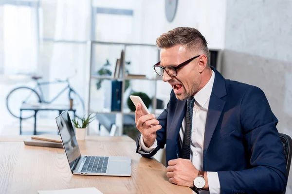 Businessman Glasses Suit Screaming While Holding Smartphone Laptop Office — Stock Photo, Image