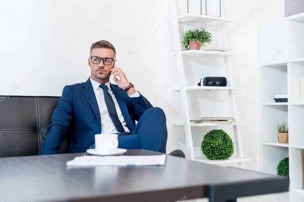 selective focus of handsome businessman in glasses talking on smartphone and sitting on sofa near coffee table 