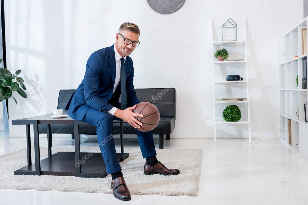 cheerful businessman in suit holding basketball in hands while sitting on coffee table