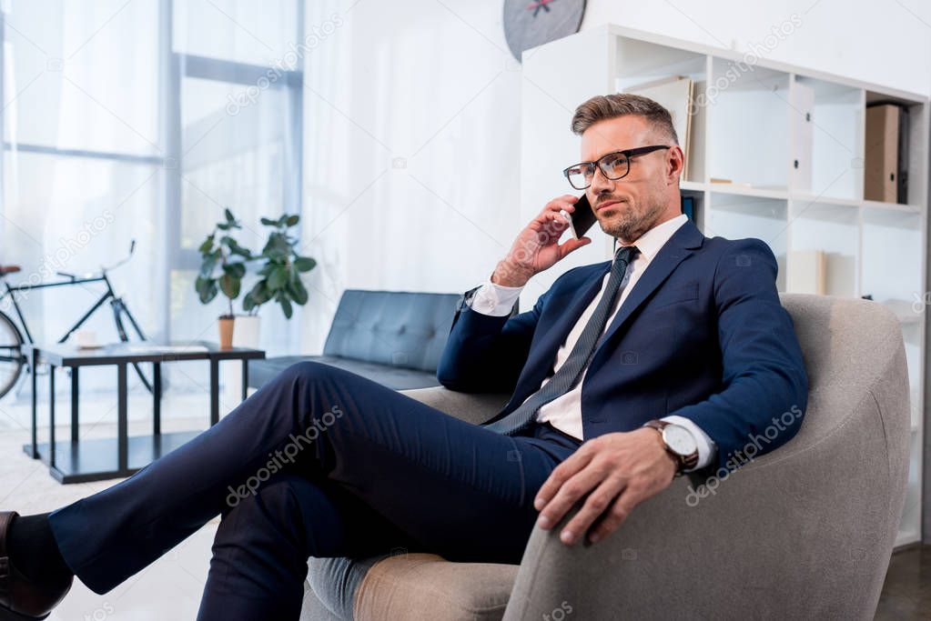 businessman in glasses sitting in armchair and talking on smartphone