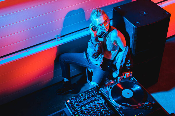 Вид на девушку-диджея со светлыми волосами, сидящую возле диджейского оборудования в ночном клубе
