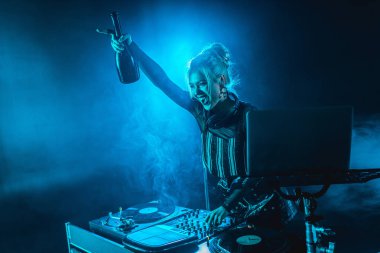 cheerful blonde dj woman in headphones holding  bottle and screaming near dj equipment in nightclub with smoke clipart