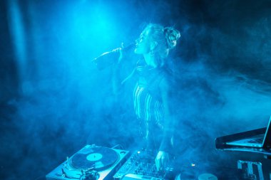 Dumanlı gece kulübünde dj ekipmanları yakınındaki şişe şarap içme Kulaklığımda mutlu sarışın dj kadın