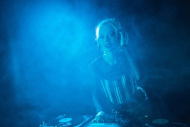 attractive dj girl listening music in headphones near dj equipment in nightclub with smoke  clipart