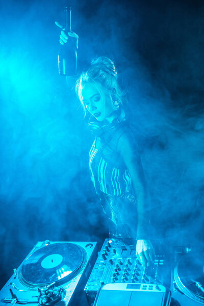 blonde dj woman in headphones holding  bottle near dj equipment in nightclub with smoke