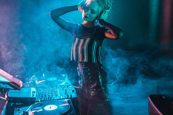 attractive dj woman with blonde hair listening music and touching headphones in nightclub with smoke