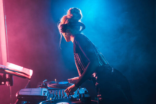 dj woman with blonde hair using dj mixer and touching vinyl record in nightclub with smoke