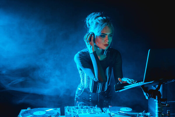 серьезная диджейка с светлыми волосами смотрит на ноутбук, слушая музыку в наушниках в ночном клубе с дымом
  