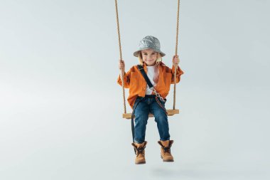 cute kid in silver hat, jeans and orange shirt sitting on swing on grey background  clipart
