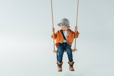 cute kid in jeans and orange shirt sitting on swing and looking away on grey background clipart