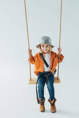 smiling kid in jeans and orange shirt sitting on swing on grey background clipart
