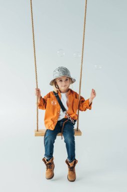 cute kid in jeans and orange shirt sitting on swing and looking at soap bubbles clipart