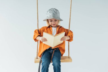 çok güzel çocuk kot pantolon ve salıncakta oturan ve gri izole kitap okuma turuncu gömlek