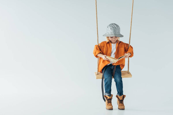 cute kid in jeans and orange shirt sitting on swing and reading book on grey background