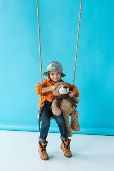 cute kid sitting on swing and holding teddy bear on blue background 