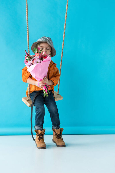 cute kid sitting on swing and holding bouquet on blue background 