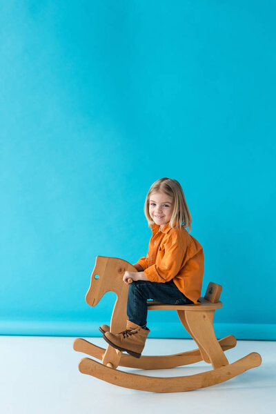 blonde and cute kid sitting on rocking horse and looking at camera on blue background 