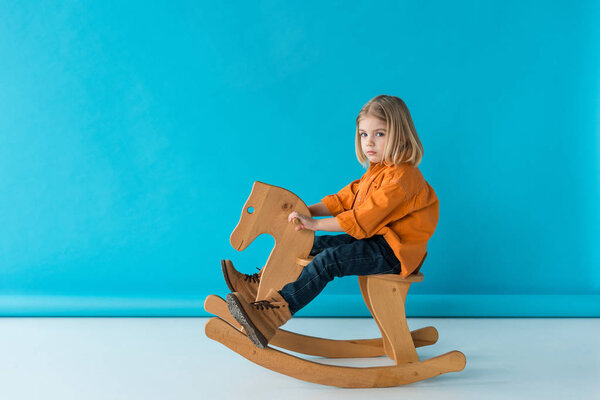 serious and cute kid sitting on rocking horse and looking at camera on blue background 
