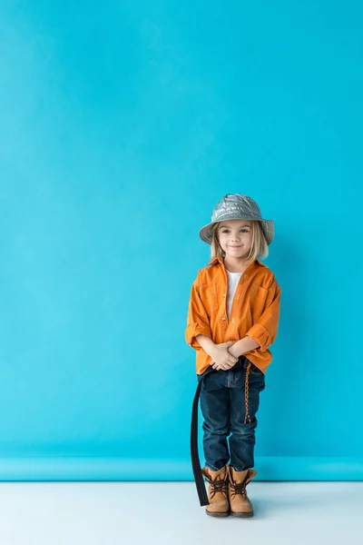銀の帽子 ジーンズとオレンジ色のシャツ 青の背景によそ見のかわいい子供 — ストック写真